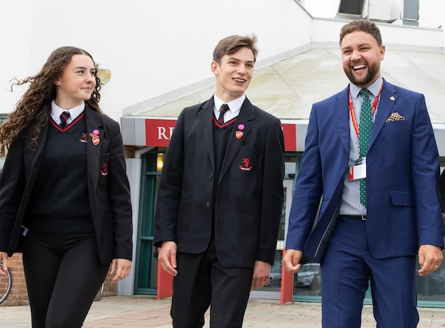 A group of students in school uniforms walking alongside an adult in a suit outside a school building.